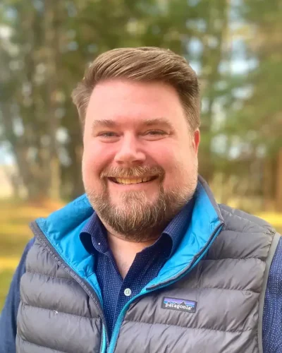 Male smiling outdoors in a blue vest and shirt, natural background, promoting mental health and well.