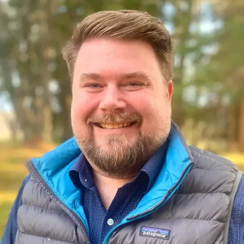 Male smiling outdoors in a blue vest and shirt, natural background, promoting mental health and well.