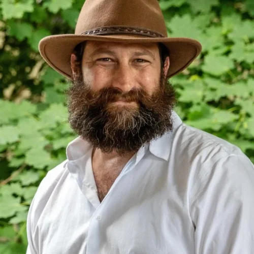 Man with beard wearing a hat outdoors, smiling, surrounded by green leaves.